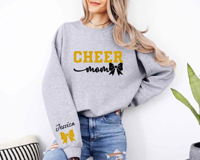 May include: Gray sweatshirt with the text "CHEER mom" in gold letters and a black bow. The left sleeve has the name "Jessica" in black letters and a gold bow.