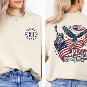 Pode incluir: Camiseta creme com design patri&oacute;tico. A frente exibe um emblema circular com o texto "1776 2026". A parte de tr&aacute;s apresenta a Est&aacute;tua da Liberdade, uma &aacute;guia e a bandeira americana, com o texto "250th Anniversary".