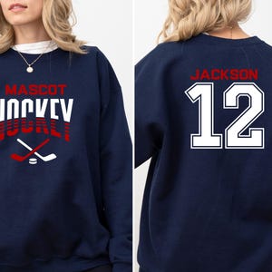 K&ouml;nnte beinhalten: Ein marineblauer Sweatshirt mit dem Text "MASCOT HOCKEY" in Rot und Wei&szlig; auf der Vorderseite und dem Text "JACKSON 12" in Wei&szlig; auf der R&uuml;ckseite.