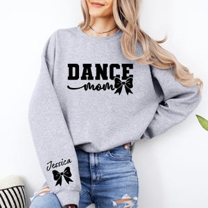 Pu&ograve; includere: Felpa grigia con il testo "Dance Mom" e un fiocco nero. La manica sinistra ha il nome "Jessica" e un fiocco nero.