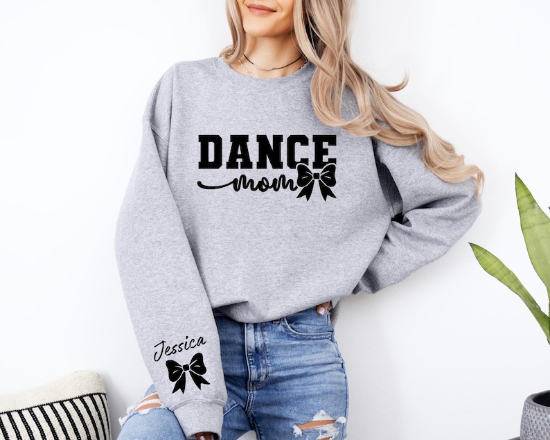 Pu&ograve; includere: Felpa grigia con il testo "Dance Mom" e un fiocco nero. La manica sinistra ha il nome "Jessica" e un fiocco nero.