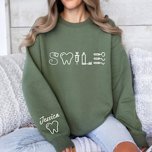 May include: A green sweatshirt with a white graphic of a tooth, syringe, and other dental tools. The text "Smile" is above the graphic. The text "Jessica" is on the left sleeve with a tooth graphic.