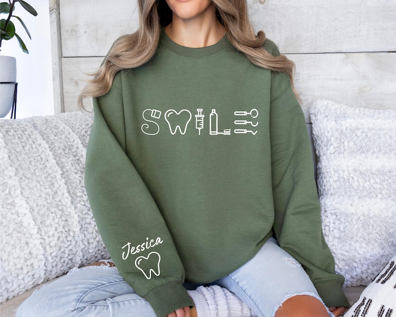May include: A green sweatshirt with a white graphic of a tooth, syringe, and other dental tools. The text "Smile" is above the graphic. The text "Jessica" is on the left sleeve with a tooth graphic.