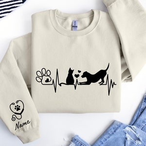May include: A beige sweatshirt with a black graphic of a cat and dog with a heartbeat line and a paw print. The text "Name" is on the sleeve.