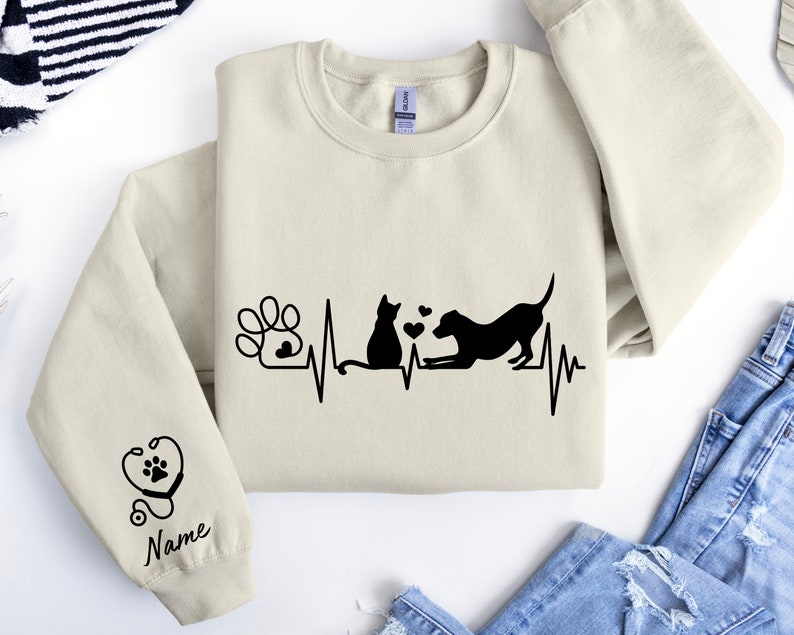 May include: A beige sweatshirt with a black graphic of a cat and dog with a heartbeat line and a paw print. The text "Name" is on the sleeve.