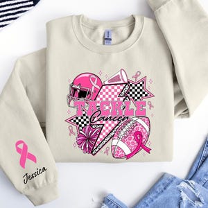 Op de afbeelding: Een beige sweatshirt met een roze en zwarte afbeelding met de tekst "Tackle Cancer". De afbeelding bevat een footballhelm, een voetbal, sterren en linten. De linkermouw heeft een roze lint en de naam "Jessica".
