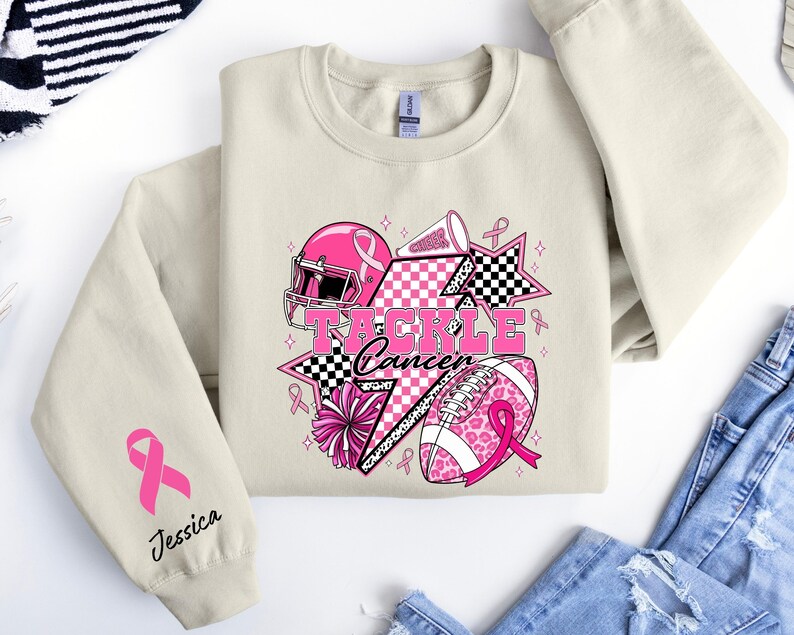 Op de afbeelding: Een beige sweatshirt met een roze en zwarte afbeelding met de tekst "Tackle Cancer". De afbeelding bevat een footballhelm, een voetbal, sterren en linten. De linkermouw heeft een roze lint en de naam "Jessica".