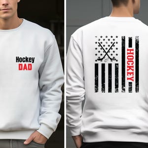 May include: White sweatshirt with a black and red graphic of a hockey stick and puck on a distressed American flag design. The text "HOCKEY" is written vertically on the right side of the flag. The front of the sweatshirt has the text "Hockey DAD" in black and red.