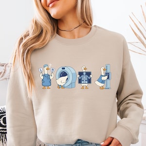 May include: Beige sweatshirt featuring a whimsical medical theme. The design includes cartoon ducks in medical roles, such as holding X-rays and operating an MRI machine. The ducks are wearing blue outfits. The letters "L" and "R" are also visible.