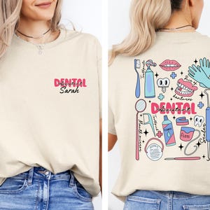 May include: A beige t-shirt with a pink and white design on the back. The design includes a variety of dental tools and the text "DENTAL ASSISTANT". The front of the shirt has the text "DENTAL Sarah" in pink.