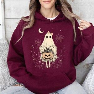May include: A maroon hoodie with a white ghost holding a jack-o-lantern. The ghost is wearing a pink bow and is surrounded by spiderwebs and flowers.