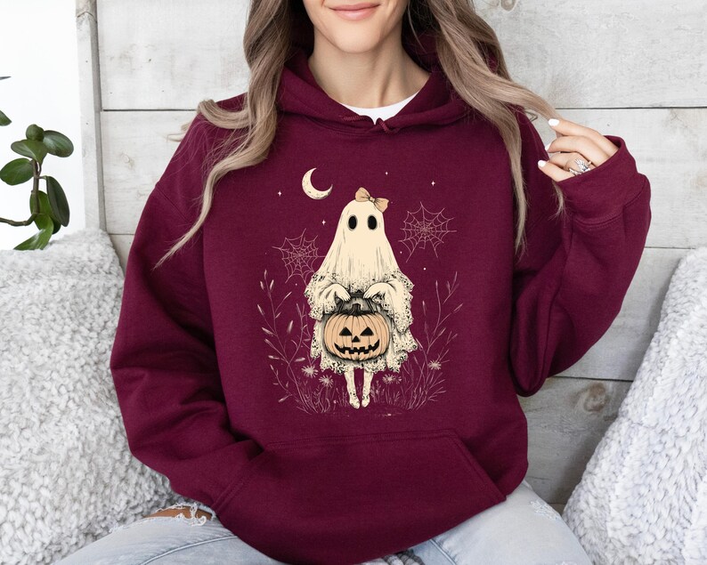 May include: A maroon hoodie with a white ghost holding a jack-o-lantern. The ghost is wearing a pink bow and is surrounded by spiderwebs and flowers.