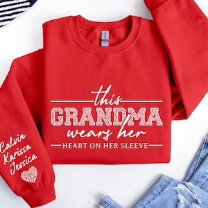 May include: Red sweatshirt with the text "This Grandma wears her heart on her sleeve" in white. The left sleeve has the names "Calvin, Karissa, Jessica" written in white.