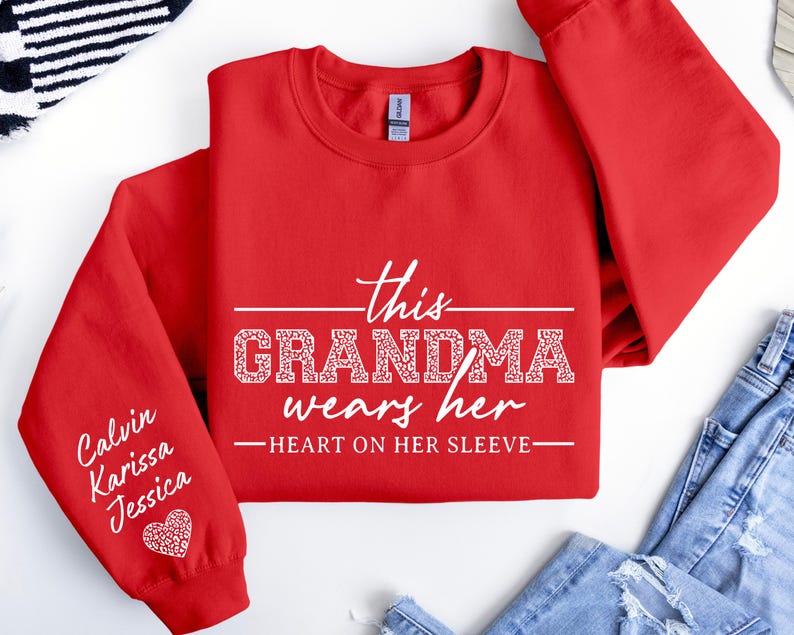 May include: Red sweatshirt with the text "This Grandma wears her heart on her sleeve" in white. The left sleeve has the names "Calvin, Karissa, Jessica" written in white.