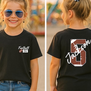 May include: A black short-sleeved t-shirt featuring a small graphic on the front chest with the text Football SIS and a football icon. The back of the shirt displays a large number 5 with a football texture and the name Jackson written across it.