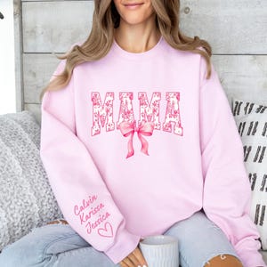 May include: Pink sweatshirt with the word "MAMA" in pink floral lettering with a pink bow. The sweatshirt has the names "Calvin, Karissa, Jessica" written on the sleeve.