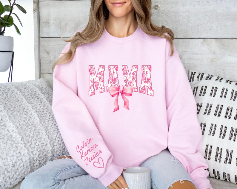 May include: Pink sweatshirt with the word "MAMA" in pink floral lettering with a pink bow. The sweatshirt has the names "Calvin, Karissa, Jessica" written on the sleeve.