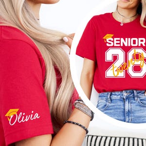 May include: Red t-shirt with the text "SENIOR 26 Twenty" in white and yellow, and a graduation cap graphic. The sleeve features the name "Olivia" in white script with a graduation cap icon. The shirt is paired with blue jeans.