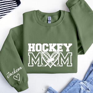 Pu&ograve; includere: Una felpa verde con il testo "HOCKEY MOM" in bianco con un cuore a forma di bastoni da hockey e un disco. La manica sinistra ha il nome "Jackson" scritto in bianco con un cuore.