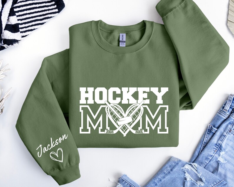 Pu&ograve; includere: Una felpa verde con il testo "HOCKEY MOM" in bianco con un cuore a forma di bastoni da hockey e un disco. La manica sinistra ha il nome "Jackson" scritto in bianco con un cuore.