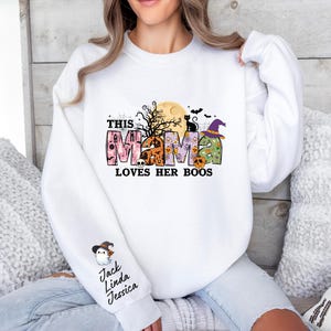 May include: White sweatshirt with a Halloween-themed design that says "This Mama Loves Her Boos". The design features a black cat, a witch's hat, and a moon with a spooky tree. The sweatshirt has a snowman graphic on the sleeve with the names "Jack, Linda, Jessica" written below.