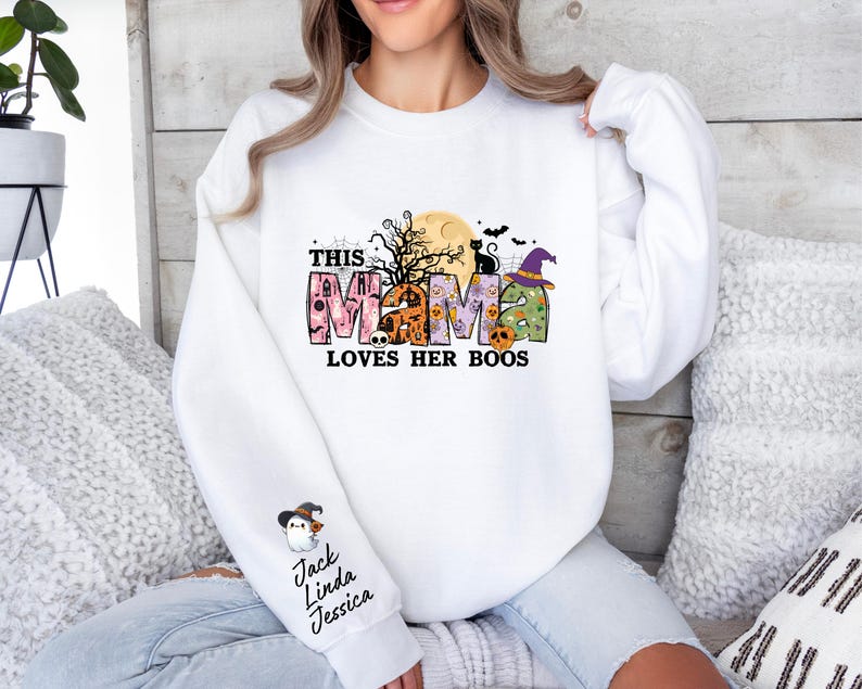 May include: White sweatshirt with a Halloween-themed design that says "This Mama Loves Her Boos". The design features a black cat, a witch's hat, and a moon with a spooky tree. The sweatshirt has a snowman graphic on the sleeve with the names "Jack, Linda, Jessica" written below.