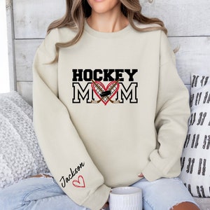 Pu&ograve; includere: Una felpa color crema con il testo "HOCKEY MOM" in nero con un cuore rosso e bastoni da hockey incrociati. Il testo "Jackson" &egrave; scritto in nero con un cuore rosso sulla manica.