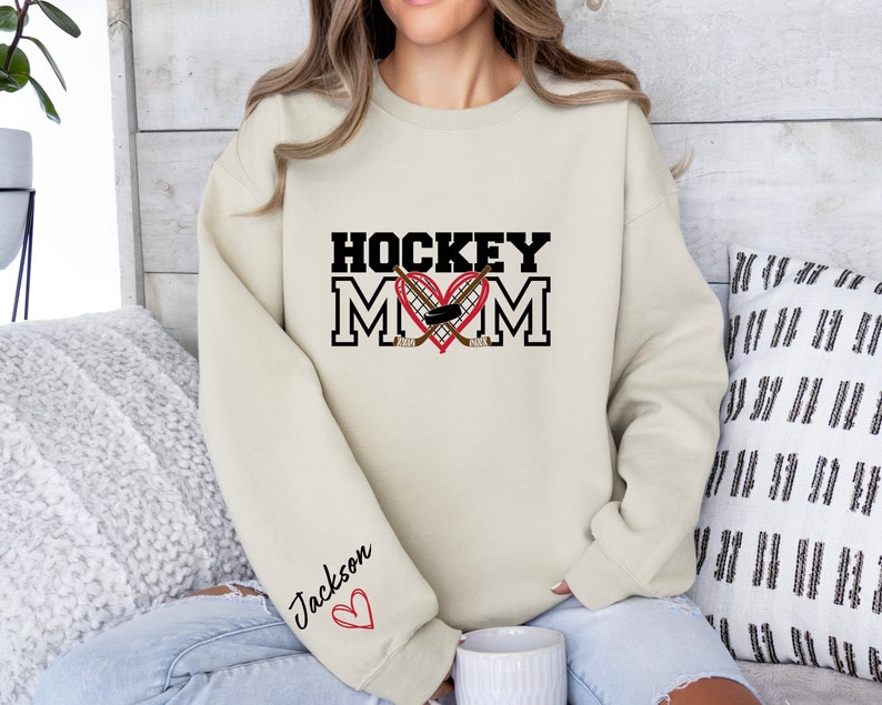 Pu&ograve; includere: Una felpa color crema con il testo "HOCKEY MOM" in nero con un cuore rosso e bastoni da hockey incrociati. Il testo "Jackson" &egrave; scritto in nero con un cuore rosso sulla manica.