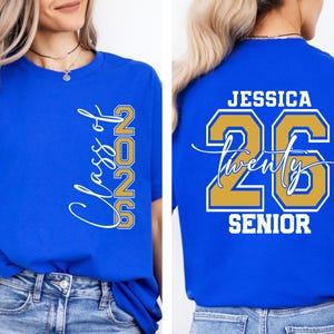 May include: Royal blue t-shirt with the text "Class of 2026" in white script and gold block letters. The back of the shirt features the name "Jessica", the number "26", and the word "Senior" in white and gold.