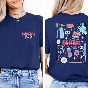 May include: A navy blue t-shirt with a pink and blue graphic design of dental tools and the text "Dental Assistant". The shirt also has the text "Dental Assistant Sarah" in pink.