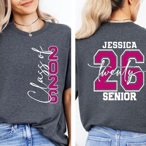 May include: Charcoal gray t-shirt with the text "Class of 2025" in white script and magenta block letters. The back of the shirt features the name "Jessica", the number "26", the word "twenty" in script, and the word "SENIOR" in white.