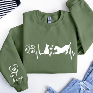 May include: Green sweatshirt with a white graphic of a cat and dog with a heartbeat line and paw print. The left sleeve has a white stethoscope with a paw print and the word "Name" underneath.