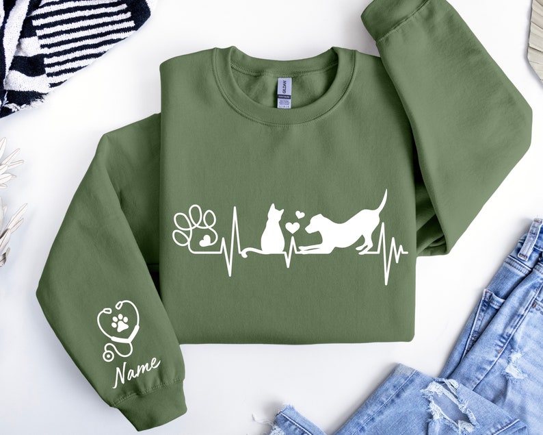 May include: Green sweatshirt with a white graphic of a cat and dog with a heartbeat line and paw print. The left sleeve has a white stethoscope with a paw print and the word "Name" underneath.