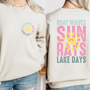 May include: A beige sweatshirt with a sun graphic and the text "Boat Waves Sun Rays Lake Days" on the front and back. The sun graphic is yellow with a pink outline and the text is in pink, blue, and yellow.