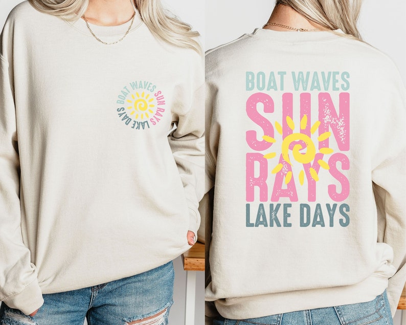 May include: A beige sweatshirt with a sun graphic and the text "Boat Waves Sun Rays Lake Days" on the front and back. The sun graphic is yellow with a pink outline and the text is in pink, blue, and yellow.