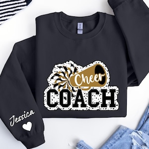 May include: Black sweatshirt with a white and gold design of a cheerleader megaphone with the text "Cheer Coach" and a white heart with the name "Jessica" on the sleeve.