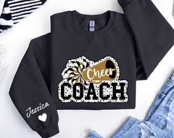 May include: Black sweatshirt with a white and gold design of a cheerleader megaphone with the text "Cheer Coach" and a white heart with the name "Jessica" on the sleeve.