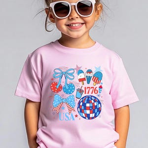 Puede incluir: Una camiseta rosa con un dise&ntilde;o patri&oacute;tico. El dise&ntilde;o incluye lazos, helados, una bola de discoteca y estrellas en colores rojo, blanco y azul. Tambi&eacute;n se incluye el texto "USA" y "1776".