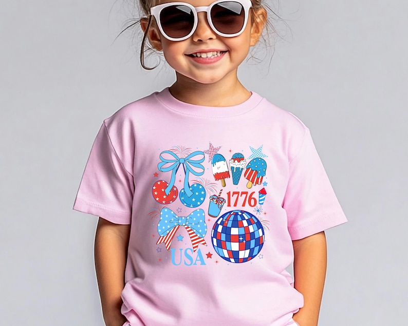 Puede incluir: Una camiseta rosa con un dise&ntilde;o patri&oacute;tico. El dise&ntilde;o incluye lazos, helados, una bola de discoteca y estrellas en colores rojo, blanco y azul. Tambi&eacute;n se incluye el texto "USA" y "1776".