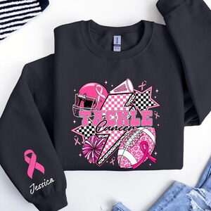 Op de afbeelding: Zwart sweatshirt met een roze en wit grafisch ontwerp met een footballhelm, football, sterren en de woorden "Tackle Cancer". De mouw heeft een roze lint en de naam "Jessica".