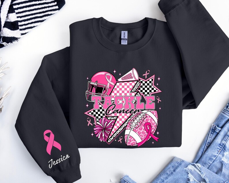 Op de afbeelding: Zwart sweatshirt met een roze en wit grafisch ontwerp met een footballhelm, football, sterren en de woorden "Tackle Cancer". De mouw heeft een roze lint en de naam "Jessica".