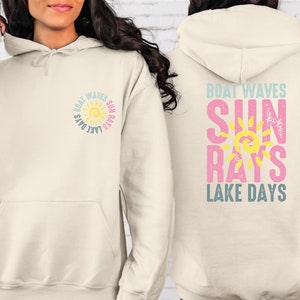 May include: A cream-colored hoodie with a sun graphic and the text "Boat Waves Sun Rays Lake Days" on the front and back. The sun graphic is yellow with orange rays and a blue outline. The text is in a blue, pink, and yellow color scheme.