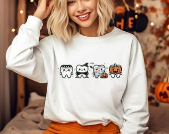 May include: White sweatshirt featuring four cartoon teeth dressed in Halloween costumes. One tooth is a ghost, another a vampire, a third is wrapped as a mummy, and the last is a pumpkin. The design is in black, orange, and white.
