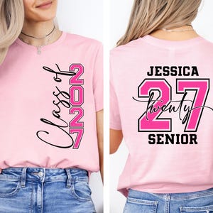 May include: A light pink t-shirt with the text "Class of 2027" in black script and pink block letters. The back of the shirt features the name "Jessica", the number "27", and the word "Senior".