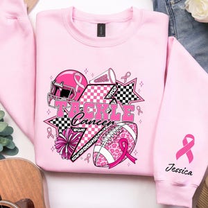 Op de afbeelding: Zachtroze sweatshirt met een grafisch ontwerp met een footballhelm, football en cheerleading-elementen. De tekst "Tackle Cancer" is prominent aanwezig. Een roze lint staat op de mouw met de naam "Jessica".
