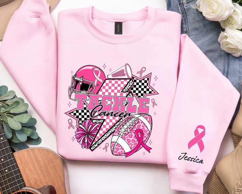 Op de afbeelding: Zachtroze sweatshirt met een grafisch ontwerp met een footballhelm, football en cheerleading-elementen. De tekst "Tackle Cancer" is prominent aanwezig. Een roze lint staat op de mouw met de naam "Jessica".