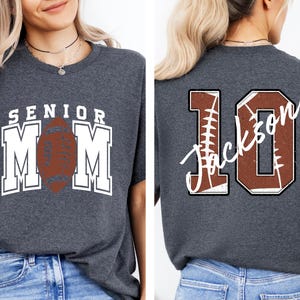 May include: A dark heather gray t-shirt featuring a front graphic with the text SENIOR MOM and a brown football. The back graphic displays the number 10 with a football texture and the name Jackson written in white cursive script across the number.