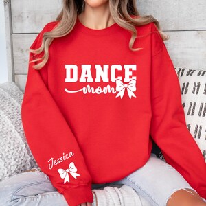 Pu&ograve; includere: Felpa rossa con il testo "Dance Mom" in bianco con un fiocco bianco. La manica sinistra ha il nome "Jessica" in bianco con un fiocco bianco.