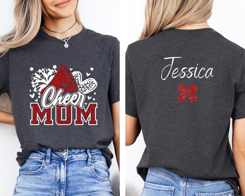 Puede incluir: Una camiseta gris oscuro con un dise&ntilde;o blanco que dice "Cheer Mom" y presenta un pomp&oacute;n de animadora rojo y blanco. La parte posterior de la camiseta tiene un lazo blanco y el nombre "Jessica" escrito en blanco.
