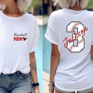 May include: White t-shirt with the text "Baseball Mom" and a heart on the front. The back of the shirt has a white baseball with a red stitching design and the number 3 with the name "Jackson" written on it.